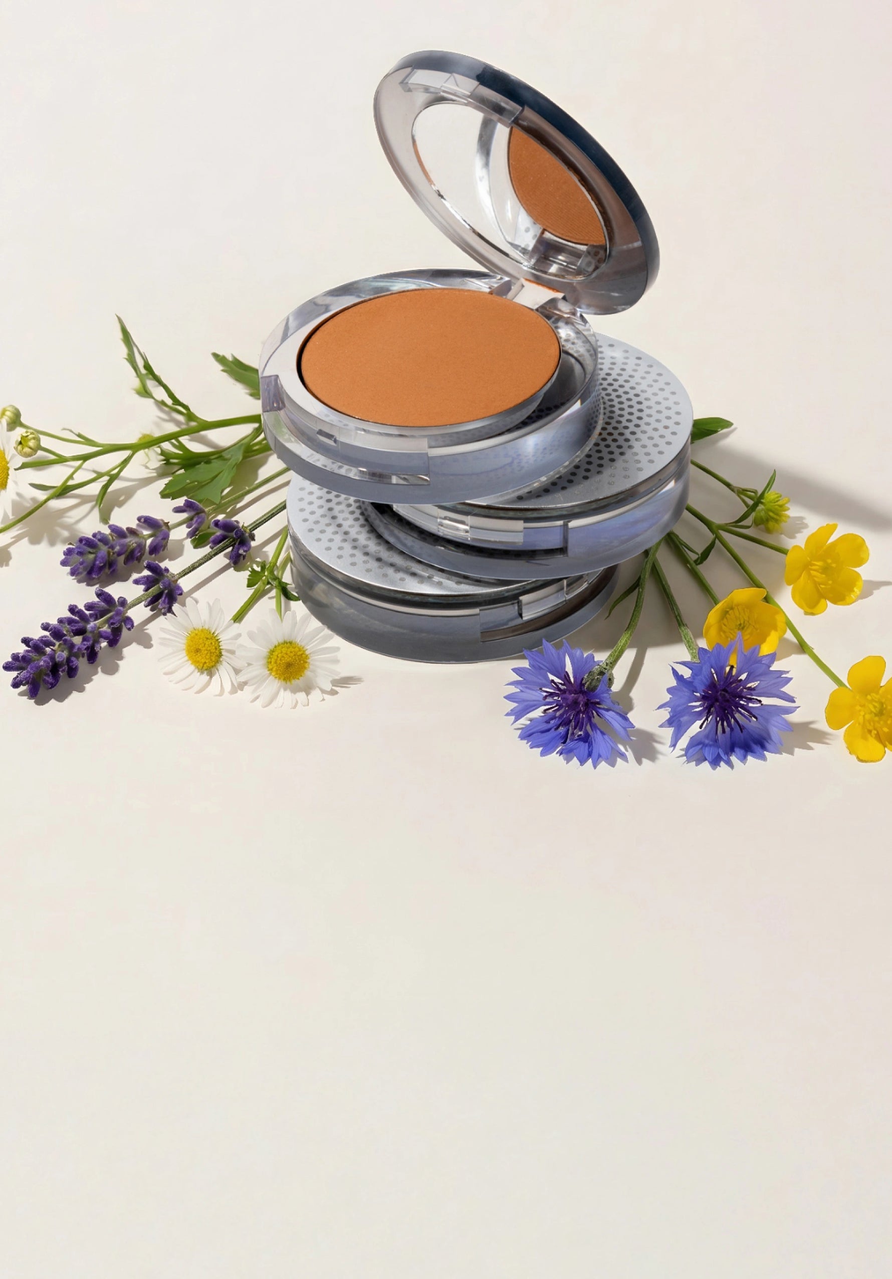 For Mom, with Love - Save up to $25: Stack of 3 4-in-1 Pressed Mineral Makeup products surrounded by yellow and purple flowers against tan background (mobile - focal point)